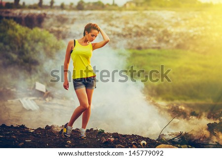 sunlight homeless young beautiful woman in a yellow tank top and denim shorts standing on a landfill in the smoke, pensive and serious Grusno