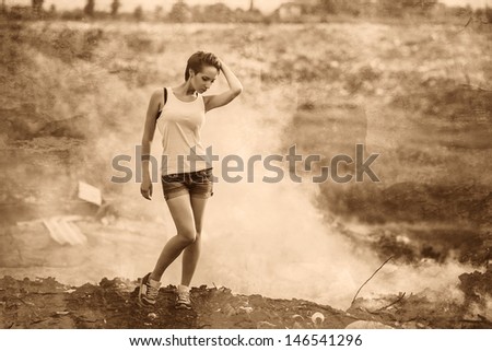 retro sepia homeless young beautiful woman in a yellow tank top and denim shorts standing on a landfill in the smoke, pensive and serious Grusno