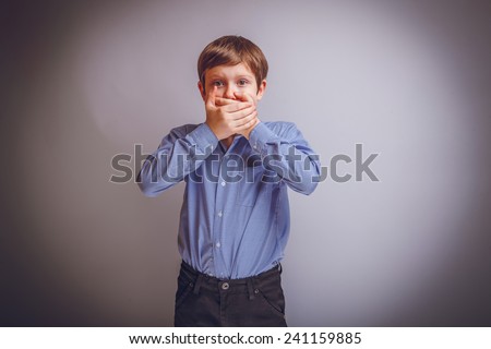 teenager boy of 10 years of European appearance surprised closed his mouth with both hands on a gray background cross process