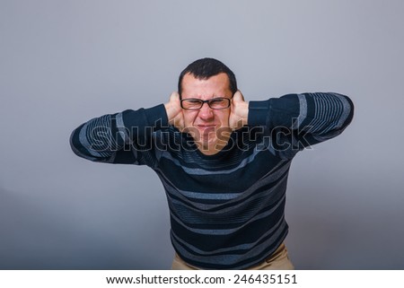 European-looking male of about thirty brunet covered his ears with his hands and closed his eyes on a gray background