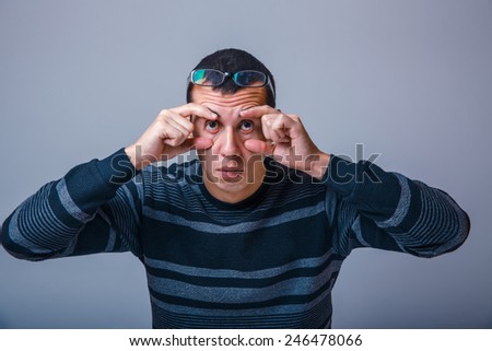 European-looking male of about thirty brunet stretches eyes with his hands on a gray background, fatigue