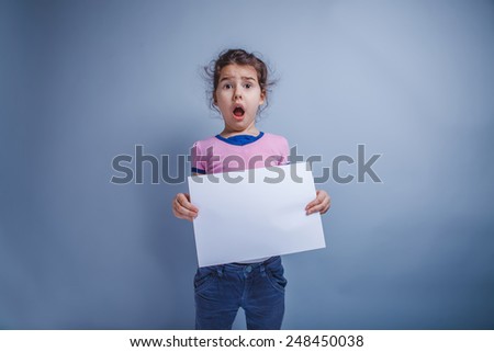 Teen girl seven years of European appearance, holding a sheet of white paper opened her mouth on a gray background, surprise