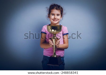 girl child 6 years of European appearance holds a cup in his hands,  happiness,  reward  and joy on a gray background shadow
