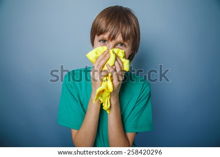 boy teenager European appearance in a green T-shirt blows his nose in a handkerchief on a gray background, flu, runny nose