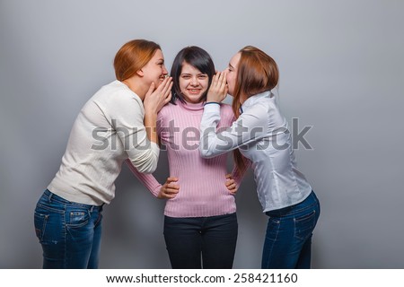 two girlfriends European appearance whisper in the ear of the third girl in a sweater on a gray background, friendship, the secret