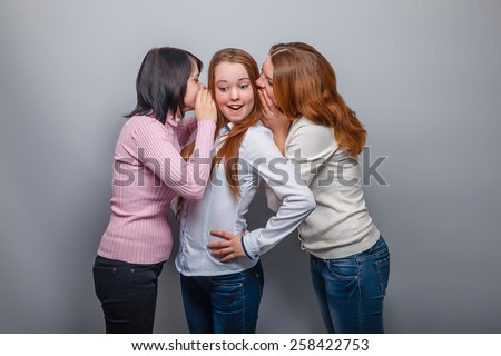 two girls European appearance blonde and brunette whispered in his ear the third blonde girl on a gray background, laughter