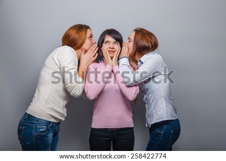 two girls in jeans European appearance whisper in the ear of the third girl in a sweater on a gray background, a secret girlfriend