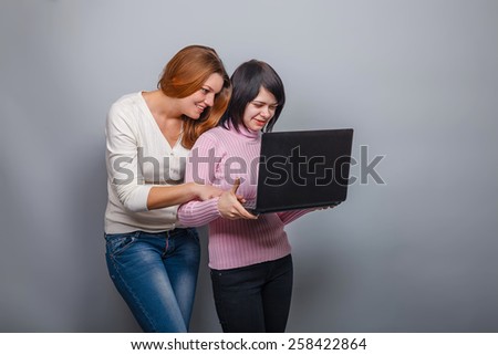 two girls European appearance blonde and brunette look into your computer and smiling on a gray background, friends, mischief