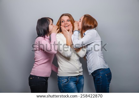 two girls European appearance whisper in the ear of the third the secret girl on a gray background, surprise