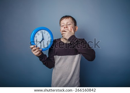 boy teenager European appearance in brown sweater holding a clock shut her mouth yawning on gray background time, drowsiness