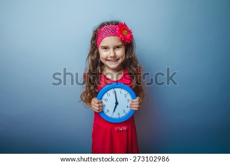 a girl of seven European appearance haired kid with a bright hairpin holding a clock on a gray background, time