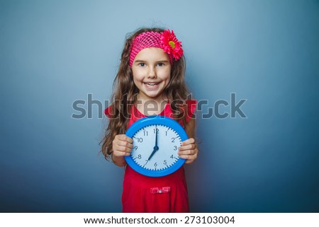 a girl of seven European appearance haired child in a bright dress holding a clock on a gray background, time, seven hours