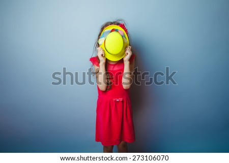 Girl European appearance haired child of seven years in a bright dress covered her face with a yellow hat on a gray background
