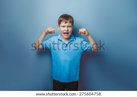 teenager boy  European appearance in a blue T-shirt shows  power of hands on a gray background, screaming, muscles