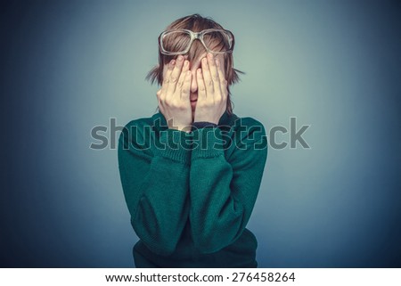 European-looking boy of ten years in glasses covered his face with his hands on a gray background instagram effect style