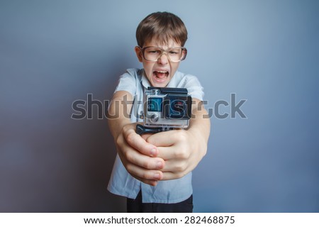 A boy of about ten European appearance in light brown shirt and glasses holding a small camera action and shouts against the gray background, video, screaming, camera gopro, go pro, action