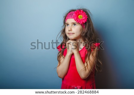 a girl of seven European appearance brunette in a bright dress folded her hands in prayer on a gray background, faith