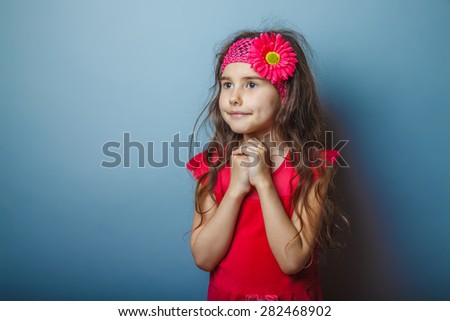 a girl of seven European appearance brunette in a bright dress folded her hands in prayer on a gray background, faith, smile