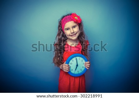 a girl of seven European appearance brunette in a bright dress holding a blue watch on gray background, time, smile retro photo effect