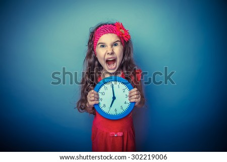a girl of seven European appearance brunette in a bright dress holding a blue clock and shouting his mouth open on a gray background, the time, the lack of retro photo effect