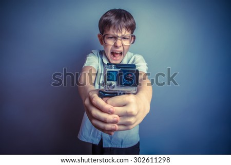 A boy of about ten European appearance in light brown shirt and glasses holding a small camera action and shouts against the gray background, video, screaming, camera retro photo effect