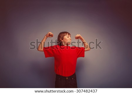 boy teenager European appearance in a red shirt shows the power of hands on a gray background retro
