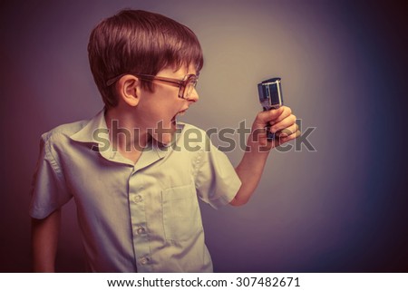 A boy of about ten European appearance in light brown shirt and glasses holding a camera action and shouts against the gray background, videography, fury retro
