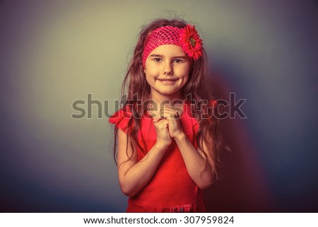 a girl of seven European appearance brunette in a bright dress folded her hands in prayer, watching in front of a gray background, smiling, portrait retro
