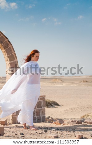 Sad wind. Sad Young redhead woman standing by an arch in a desert.