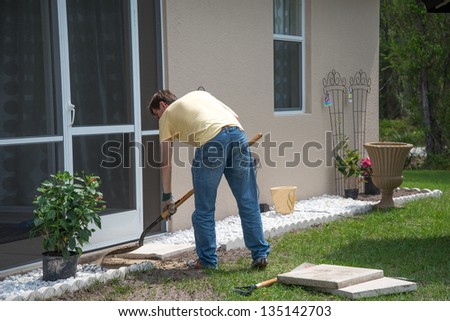 Landscaping Work - A man working outdoors.  He is using a shovel to level the dirt to place concrete patio slabs in the landscaping near the door to the house.