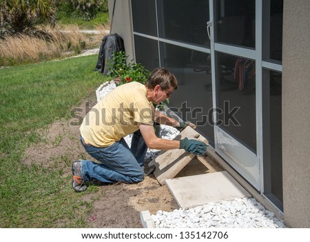 Landscaping Work - A man working outdoors.  He is placing heavy concrete patio slabs in the landscaping near the back door to the house.