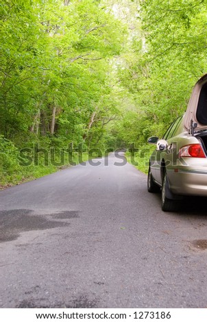 A stranded car along the side of a country road with it\'s  gas tank open.