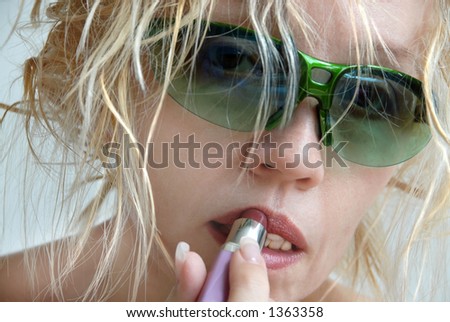 A blonde woman with messy hair, putting on her lipstick and hiding behind green sunglasses. Bad hair day.  Drugged up.