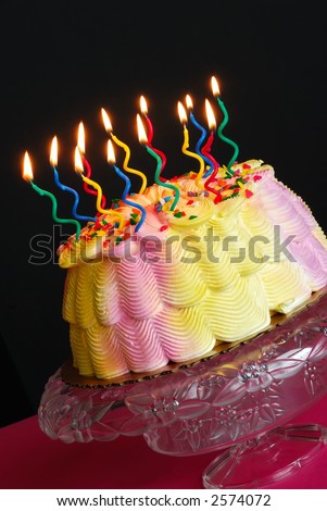 Birthday Cake With Lighted Candles - Burning candles light up a pink and yellow iced birthday cake that sits on a decorative cake plate in front of a black background.