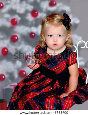 Bah Humbug ... a cute expression on the face of an 18 month old girl sitting in front of a christmas tree, holding holiday ornaments in her hand.