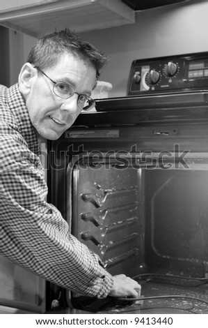 Monochrome image of a man cleaning his oven in his kitchen and looking directly at the viewer.