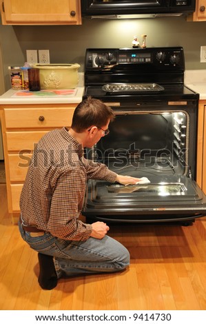 A man kneels on the floor in the kitchen and cleans the oven.  Stay at home dad.