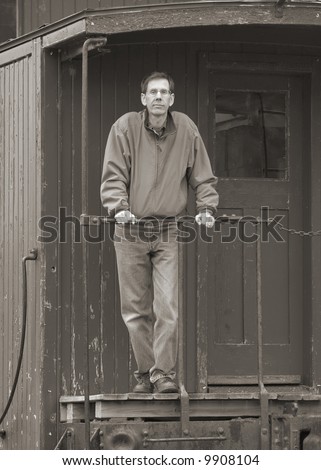Man standing on the back of an old train car caboose, leaning on the rail.