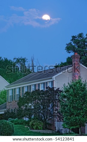 Full moon in the clouds over the rooftop of a house at dusk just before nightfall.