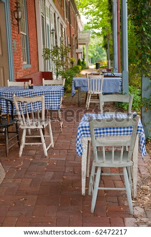 An outdoor cafe with vintage chairs, tables, blue and white checked table cloths from the 1950s, set up along the cobblestone walkway between the old buildings in a small town.