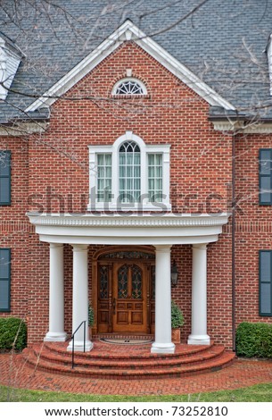 Stately white columns decorate the front entry of a brick home.  Stained glass windows in the wood door, sidelights and a transom window add to the look of luxury.