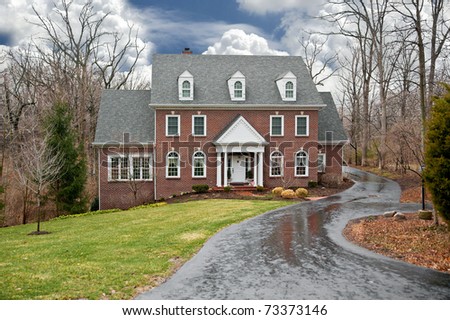 A brick two-story house in a secluded wooded setting with an asphalt driveway on a wet, rainy winter day.