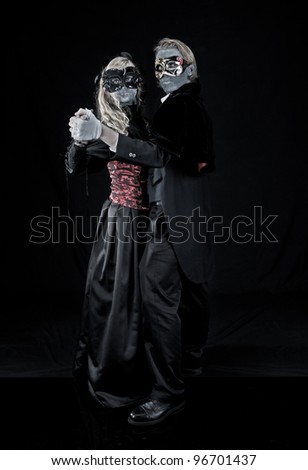 Young blonde couple dressed in vintage goth vampire style clothing poses in a dance position.  They are wearing heavy, grey colored makeup and masks.