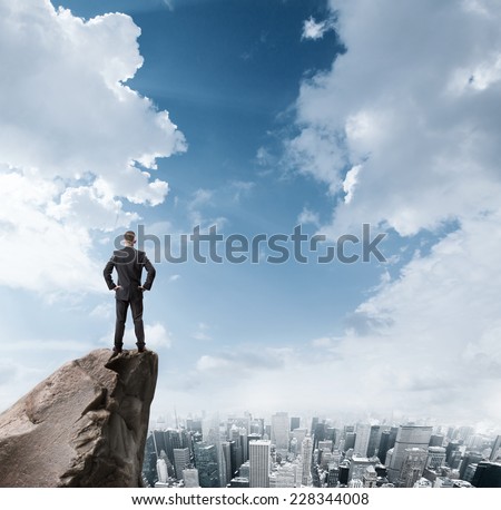 Young businessman standing on edge of rock mountain and looking at the city Young businessman standing on edge of rock mountain and looking at the city