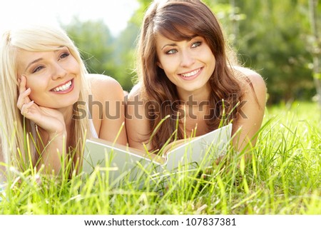 Two young beautiful smiling women reading book, sitting on grass at summer green park.