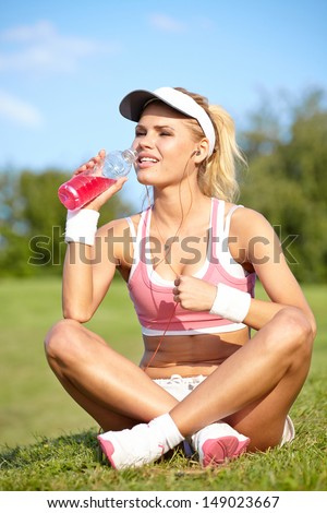 Stunning young blonde woman in pink sports bra rests while holding a water bottle and adjusting music on portable music player