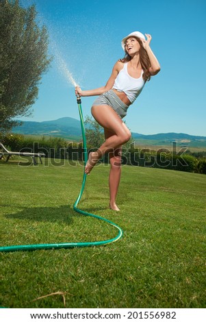 Beautiful young woman having fun in summer garden with garden hose splashing summer rain.