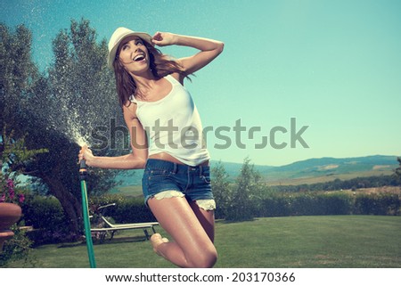 Beautiful young woman having fun in summer garden with garden hose splashing summer rain.