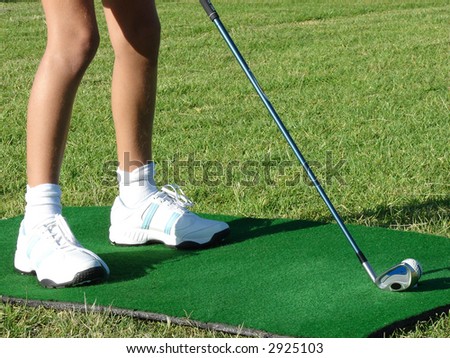 Young girl in white shoes standing on a practicing mat during golf lesson, holding a golf club