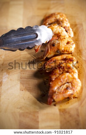 Shrimp dish, tongs pick up grilled shrimp on wood plate.